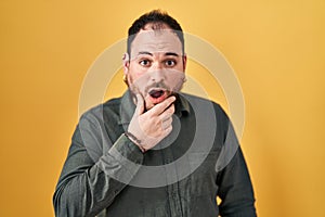 Plus size hispanic man with beard standing over yellow background looking fascinated with disbelief, surprise and amazed