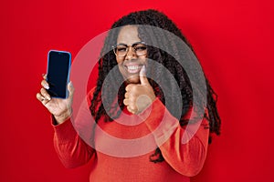 Plus size hispanic woman holding smartphone showing blank screen smiling happy and positive, thumb up doing excellent and approval