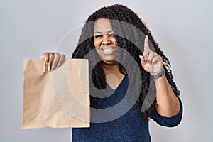 Plus size hispanic woman holding take away paper bag surprised with an idea or question pointing finger with happy face, number