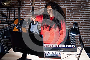 Plus size hispanic woman playing piano at music studio looking unhappy and angry showing rejection and negative with thumbs down