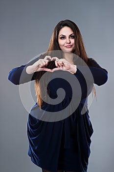 Plus size model in blue dress making a heart symbol, fat woman with long hair on gray background, body positive concept