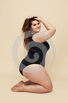 Plus Size Model. Fat Woman In Black Bodysuit Portrait. Brunette Touching Hair And Posing On Beige Background.