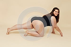 Plus Size Model. Fat Woman In Black Bodysuit Portrait. Crawling Brunette Posing On Beige Background.