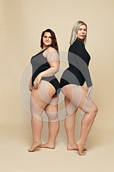 Plus Size Models. Full-figured Women In Black Bodysuits Full-length Portrait. Brunette And Blonde Posing On Beige Background.