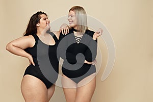 Plus Size Models. Full-figured Women Portrait. Smiling Brunette And Blonde In Black Bodysuits Posing On Beige Background.