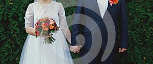 Plus size wedding couple is standing and hugging outside. Curvy bride is holding beautiful colorful bouquet with orange, red and