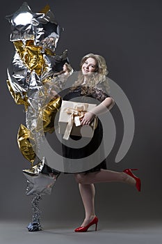 Plus size woman in black dres with gift box and balloons on gray background