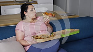 Plus size woman eats slice of pizza with great pleasure sitting on a couch