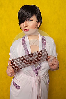 Plus size woman stands in a pink Bathrobe and holding in her hands a huge bar of chocolate on yellow background