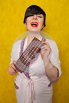 Plus size woman stands in a pink Bathrobe and holding in her hands a huge bar of chocolate on yellow background