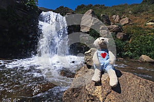 plush toy teddy bear traveler sits on rock at waterfall of river on vacation in Kaskasu gorge in summer