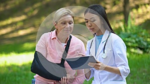 Podiatric physician with tablet and elderly woman in arm sling smiling on camera