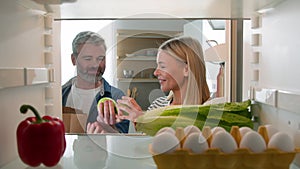 Point of view POV from inside refrigerator middle-aged couple unpack food bag in kitchen adult man and woman husband
