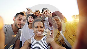 Point of view shot of African American girl holding camera and taking selfie with happy friends at party on roof. Men