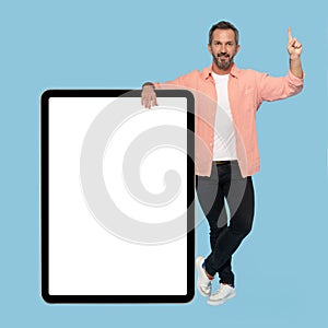 Pointing finger up middle aged grey haired man leaned on big digital tablet with white screen happy smiling on camera