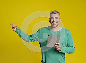 Pointing sideways smart middle aged handsome man holding digital tablet in hands smiling looking at camera wearing