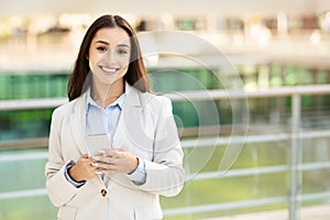 A poised young woman in a business suit smiles warmly while holding and looking at her smartphone