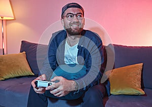 Poisitive young adult man gamer player holding gamepad controller playing video game sitting alone on sofa in the living room at