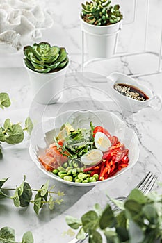 Poke bowl , buddha boul with fresh salmon,boiled eggs, avocado, cherry, bell pepper and greens at the white plate on light marble