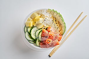 poke bowl with salmon, avocado, mango, rice, cucumbers, sprinkled with white and black sesame islated with chopsticks on white