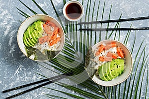 Poke bowl with salmon rice and vegetables on grey background with palm leaves. Traditional Hawaiian raw fish salad. View from