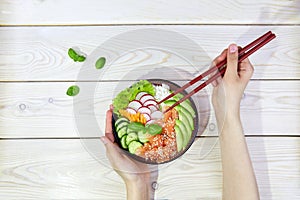 Poke bowl with salmon, rice and vegetables. Women's hands hold chopsticks and a bowl with hawaiian ahi