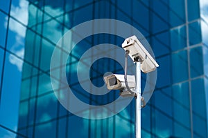 Pole with two white video surveillance cameras at big office glass building background