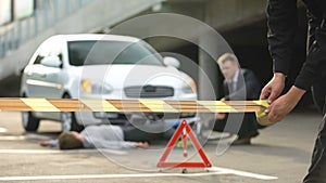 Policeman taping car accident area, male driver sitting near victim on road