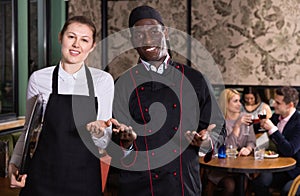 Polite waitress standing in restaurant hall with cheerful African American chef, meeting guests