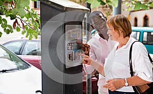 Polite intelligent African man helping middle aged woman to buy ticket in parking meter on summer city street