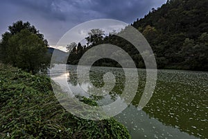 A polluted river flowing between trees and bushes under dark clouds at sunset