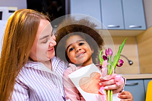 polyethnic mom and her daughter with afro hairstyle sitting in the kitchen with a bouquet of tulips and posrcard