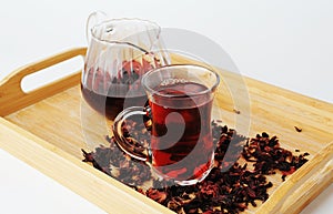 Pomegranate flowers tea in glass cup closeup. Teapot in the background. Bamboo tray with scattered petals. Ruby infusion