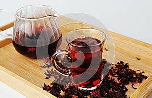 Pomegranate flowers tea in glass cup closeup. Teapot in the background. Bamboo tray with scattered petals. Ruby infusion