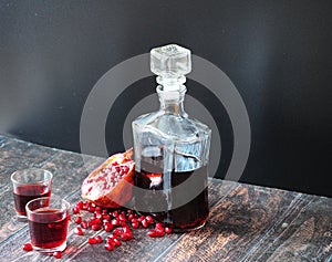 Pomegranate liqueur in a glass decanter and two glasses stands on a wooden table against a black wall, next to it is ripe broken