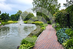 A pond filled with water with a spray jet fountain in a park with pedestrian sidewalks.