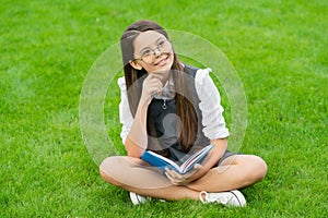 pondering child in glasses reading book sitting on green grass