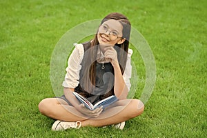 pondering child in glasses reading book sitting on green grass