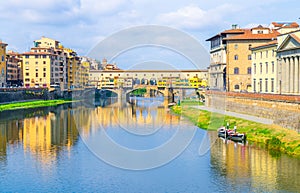 Ponte Vecchio bridge with colourful buildings houses over Arno River blue reflecting water and boats near river bank in Florence