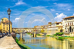 Ponte Vecchio stone bridge with colourful buildings houses over Arno River blue reflecting water and embankment promenade in histo