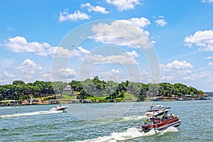 Pontoon boat full of people and two speedboats race down lake with luxury homes and docks on shore under bright blue sky with