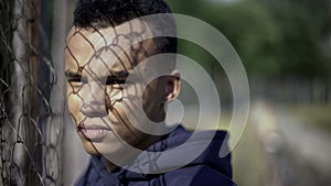 Poor afro-american teenager standing by metal fence, life difficulties concept