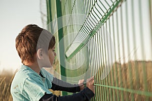 Poor dirty little caucasian boy refugee standing near state border fence holding it with hands with hope of help and