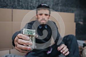 Poor and dirty man is sitting on the ground and looking on camera. He is holding a metal cup with dollar in it. He has