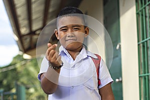 A poor but friendly rural school kid makes a finger heart gesture. Outside a simple small classroom building