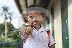 A poor but happy rural school kid pointing to the camera confidently with his finger. Outside a simple small classroom building