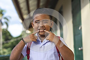 A poor but happy rural school kid shows his nice toothy smile with both fingers. Outside a simple small classroom building