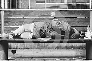 Poor homeless man or refugee sleeping on the wooden bench on the urban street in the city, social documentary concept, black and w