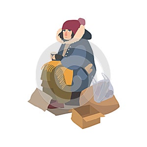 Poor homeless woman dressed in ragged outerwear sitting on street beside pile of garbage, holding paper cup and begging