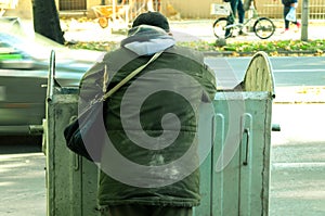 Poor and hungry homeless man in dirty clothes looking for food in the dumpster on the urban street in the city.